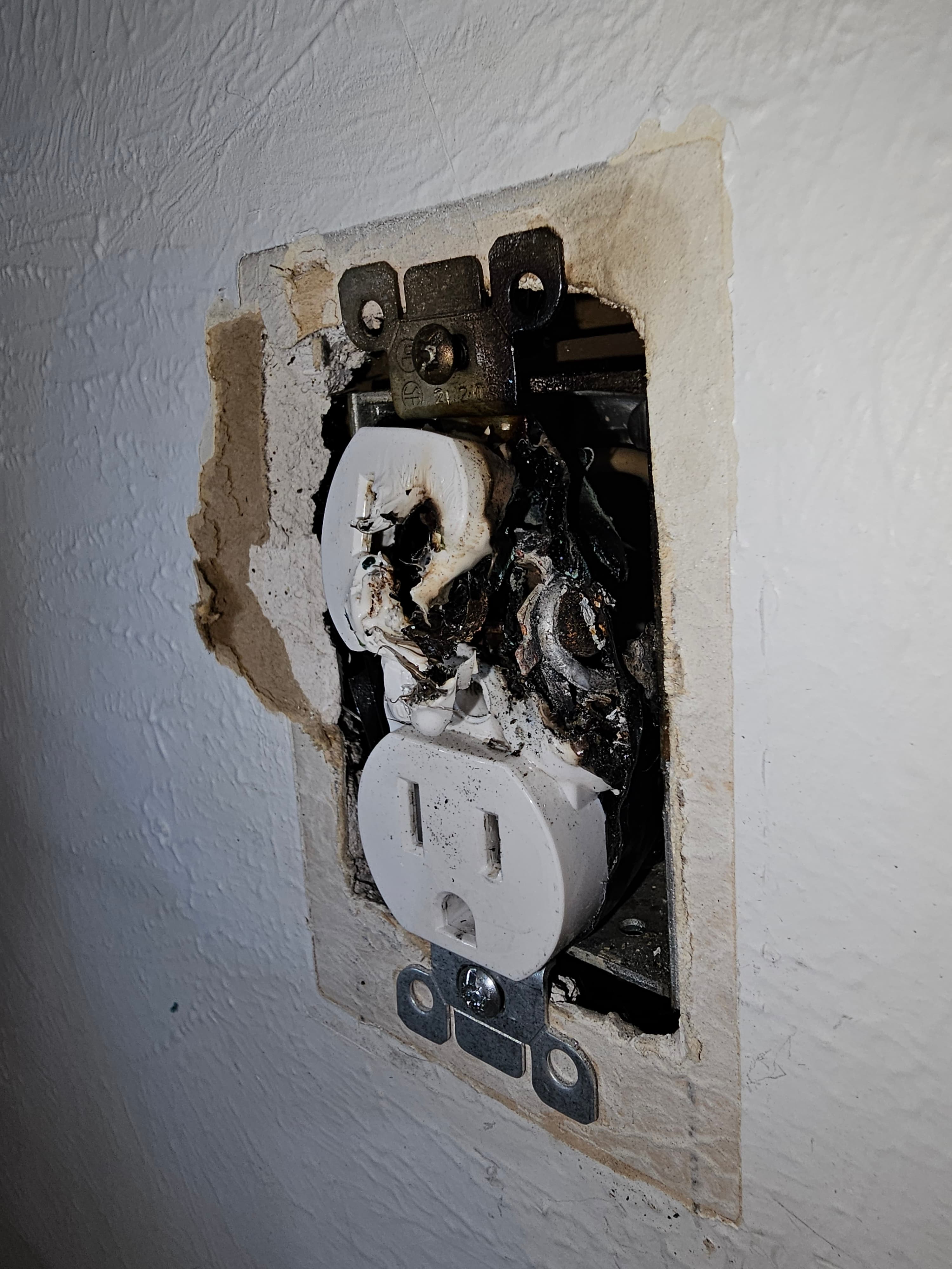Severely burned and melted white electrical outlet with fire damage in a damaged wall.