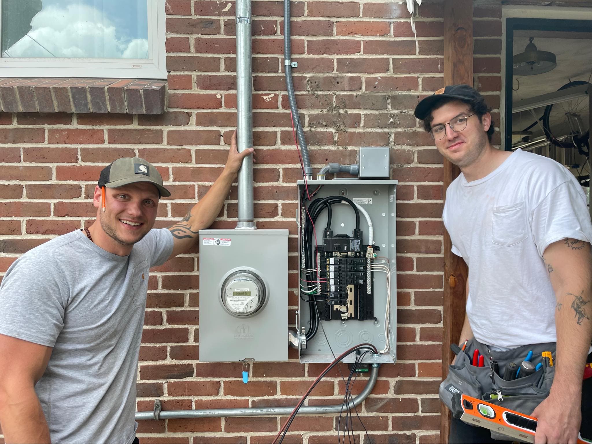 Two smiling electricians posing next to an open electrical panel and meter on a brick wall.