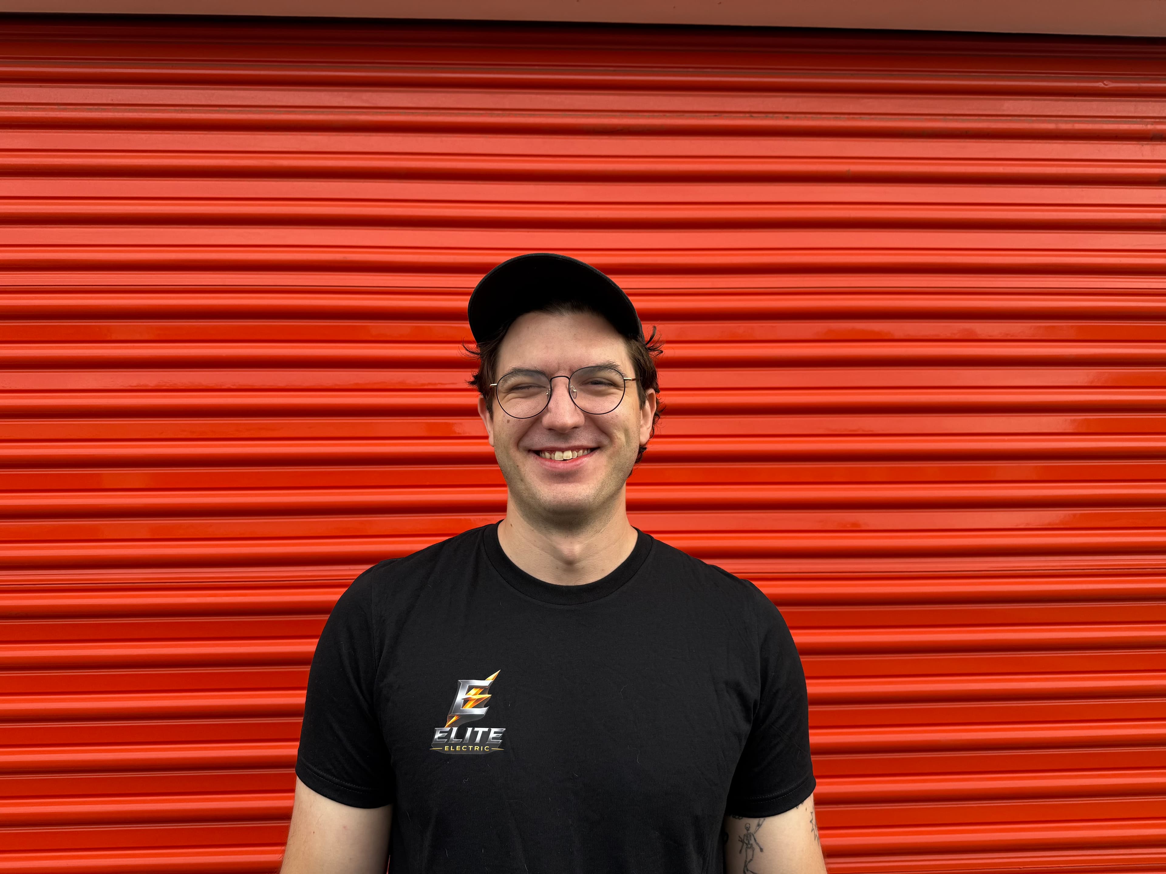 Smiling man in glasses and a black Elite Electric shirt against a red corrugated door.