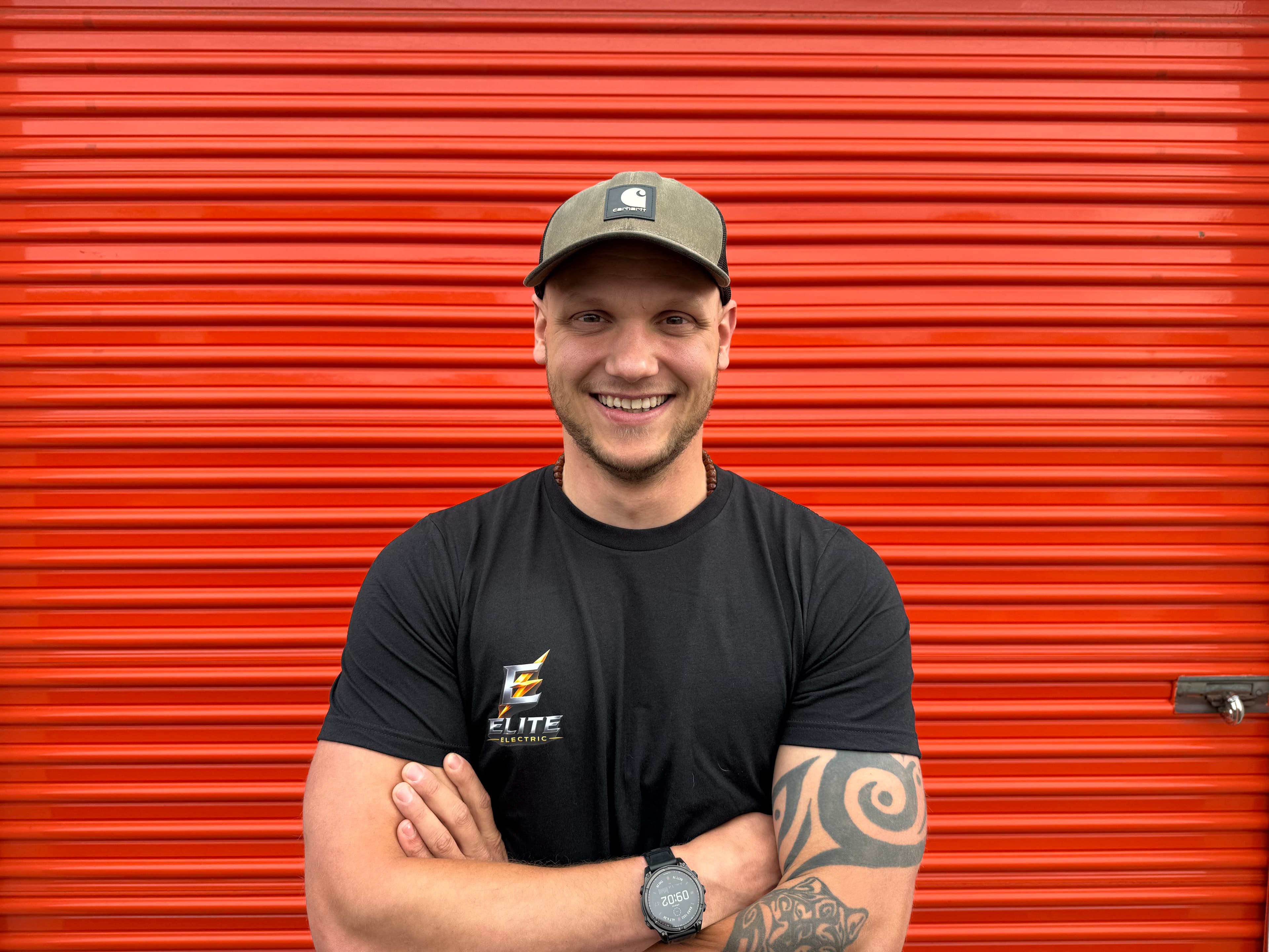 Smiling man in black t-shirt and cap with arms crossed against orange corrugated door.